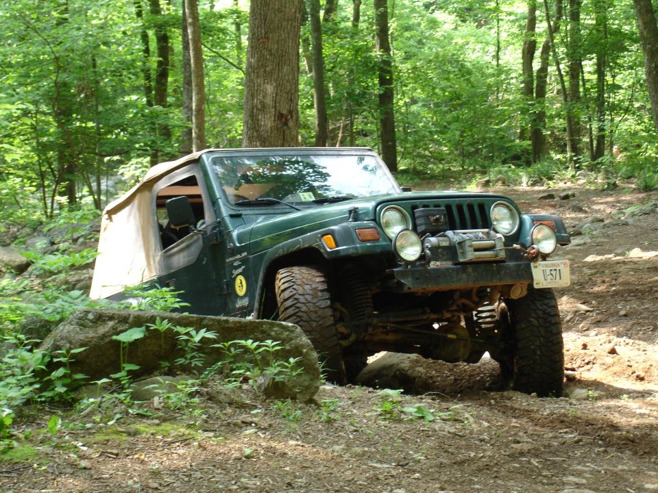 Green Jeep Wrangler with a tan soft top is navigating a rocky, wooded trail. It has a winch mounted on the front bumper and a license plate reading U-571