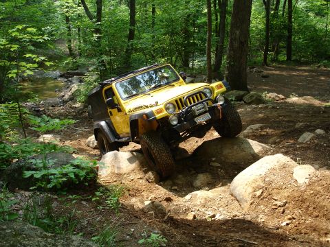 1996 Ford Bronco - A yellow Jeep Wrangler tackles a challenging off-road course with large rocks and dirt.