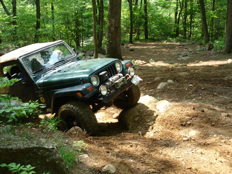 Green Jeep Wrangler navigating a rocky, wooded trail, with one front tire on a mound of dirt