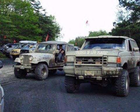 1991 Ford Bronco - Two off-road vehicles, a Ford Bronco and a Jeep CJ, are parked side-by-side on a dirt path with trees in the background.