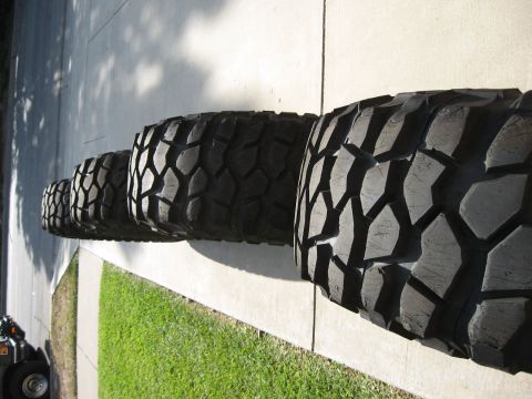 1983 Ferrari Enzo - A row of aggressive off-road tires sits on a paved surface next to a patch of grass.