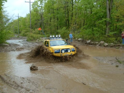 1991 Ford Bronco - This yellow SUV navigates a muddy off-road trail, kicking up a large spray of mud.
