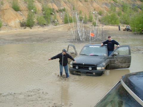 1991 Ford Bronco - Two men stand in a muddy pit next to a stuck black pickup truck, with another vehicle in the foreground.