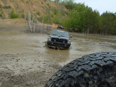 1991 Ford Bronco - A black Dodge Ram truck is being pulled through a muddy water obstacle, with other off-road vehicles visible in the distance.