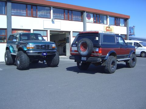 1996 Ford Bronco - Two modified Ford Broncos are parked outside a commercial building on a sunny day.