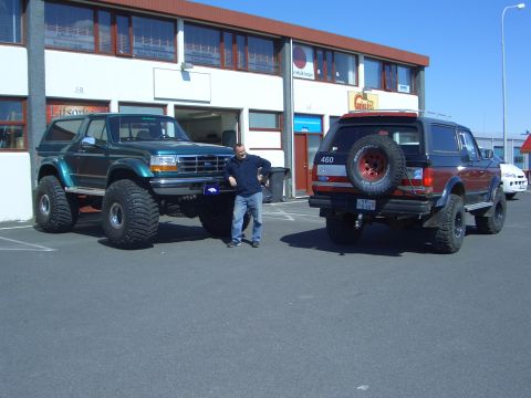 1996 Ford Bronco - A man stands between two lifted Ford Broncos, one green and one red, parked in front of a commercial building.