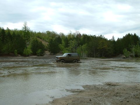 1991 Ford Bronco - A 1991 Ford Bronco navigates a challenging muddy terrain, kicking up water as it drives through the mire.