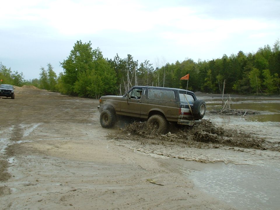 Muddy 1991 Ford Bronco driving through a dirt and water obstacle course. Another SUV is visible in the background