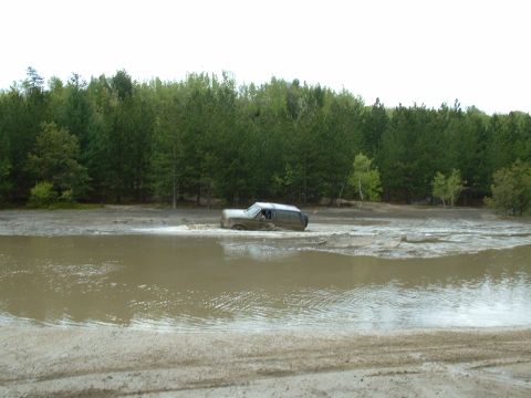 1991 Ford Bronco - An SUV navigates through a muddy water obstacle, surrounded by a dense forest.