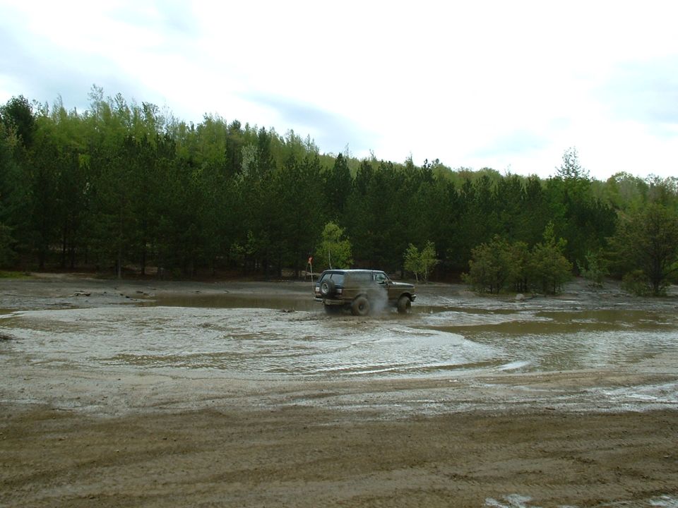 1991 Ford Bronco driving through a muddy field with trees in the background