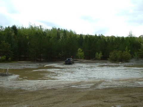 1991 Ford Bronco - A truck navigates a muddy terrain with a dense forest bordering the area.