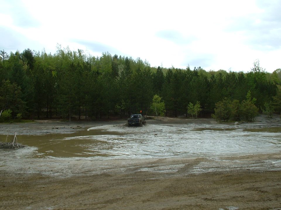 Dark colored truck driving through a muddy, wet area with trees in the background