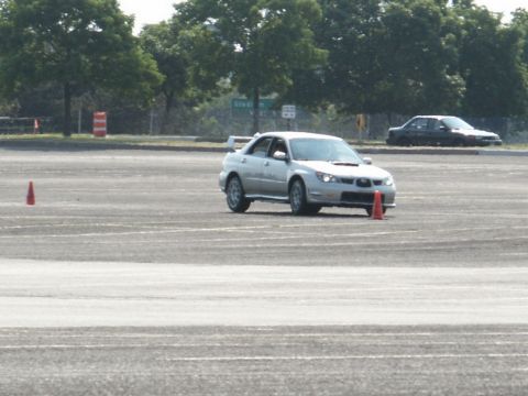 2001 Mercury Grand Marquis - A silver Subaru Impreza WRX drives on a paved lot with cones, while another car is visible in the background.