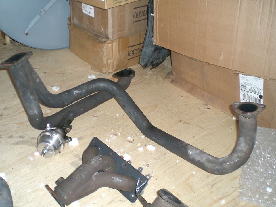 Exhaust manifold and pipes with a turbocharger wastegate, on a wooden floor with cardboard boxes in the background