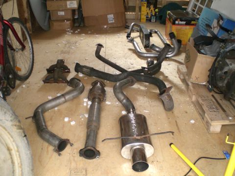 1995 Ford Probe - Exhaust system components laid out on a plywood garage floor: a polished stainless axle-back muffler with a large rolled tip, a catalytic c…