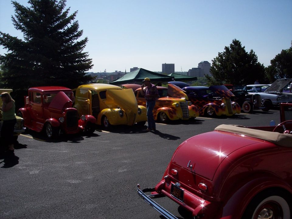 Several hot rod cars parked in a lot, some with hoods up, under a clear sky