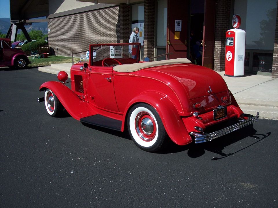 Red 1932 Ford convertible hot rod with whitewall tires parked next to a vintage gas pump. Another car is visible to the left