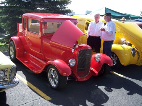 1972 Ford Ranchero - Two men admire a bright red hot rod coupe at an outdoor car show, with a yellow vehicle nearby.