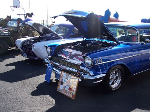 1972 Ford Ranchero - Two classic cars are displayed with their hoods open at an outdoor event, with a framed collection of images in the foreground.