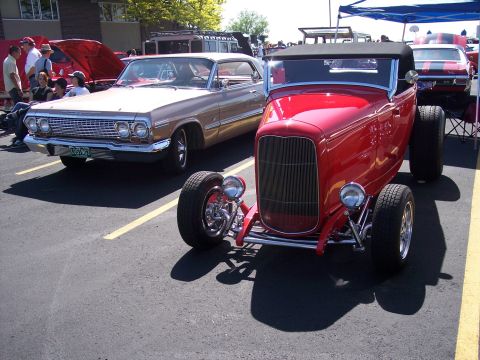 1972 Ford Ranchero - A red hot rod is parked in front of a tan 1963 Chevrolet Impala at a car show.