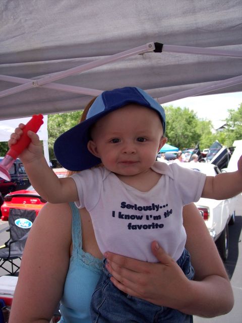 1972 Ford Ranchero - A baby wearing a blue cap and a white onesie with text is held up in front of a car show.