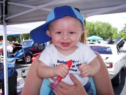 1972 Ford Ranchero - A smiling baby wearing a blue baseball cap is held up in front of a car show with several vehicles visible.
