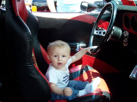 1972 Ford Ranchero - A baby sits in a car's interior, reaching for the steering wheel. The seats are black and red racing style.