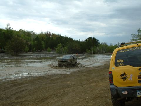 1991 Ford Bronco - A brown SUV navigates a muddy off-road course, while a yellow SUV is parked nearby.