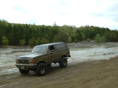 1991 Ford Bronco - 1991 Ford Bronco navigates a muddy off-road course with a forest backdrop.