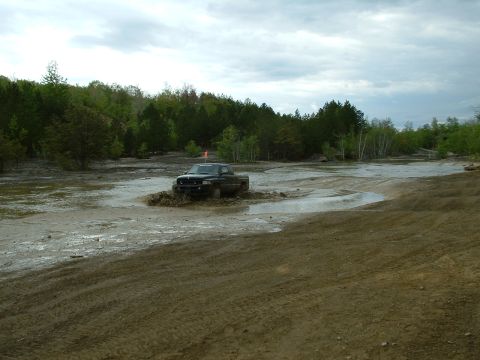 1991 Ford Bronco - A dark pickup truck navigates a muddy terrain with trees in the background under a cloudy sky.