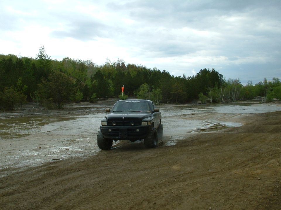 Black pickup truck with large tires driving through mud and water, trees in background