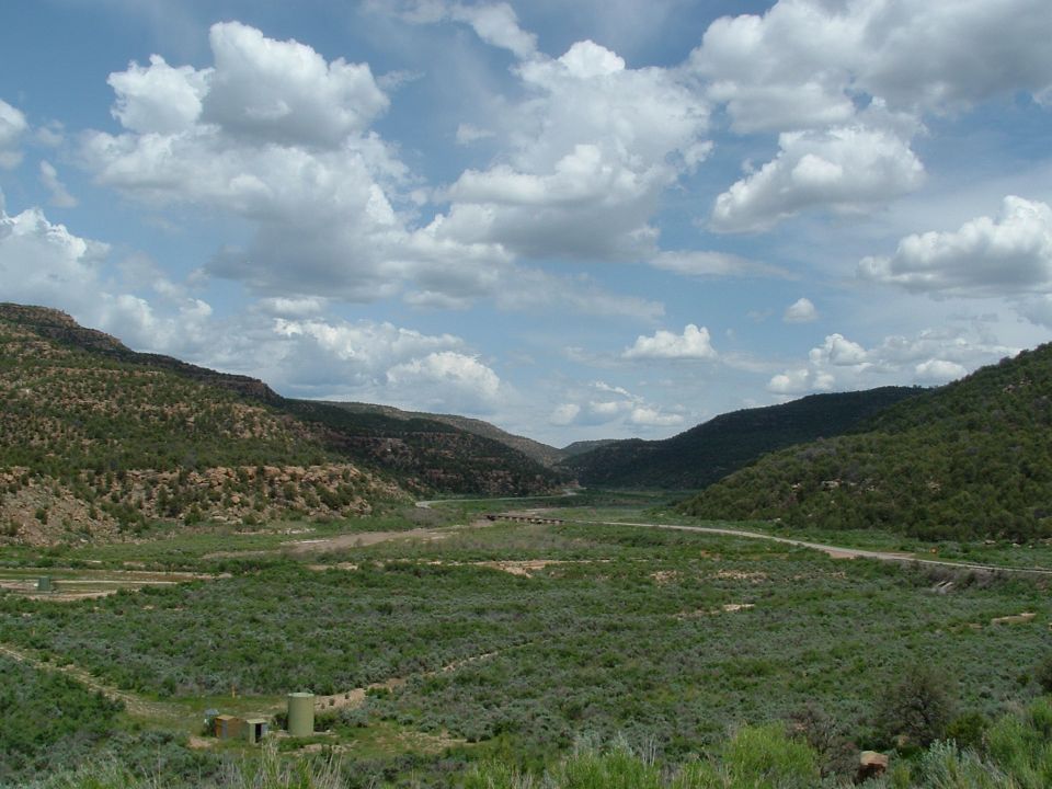 Wide shot of a valley with mountains on either side, a road winding through it, and green brush covering the valley floor
