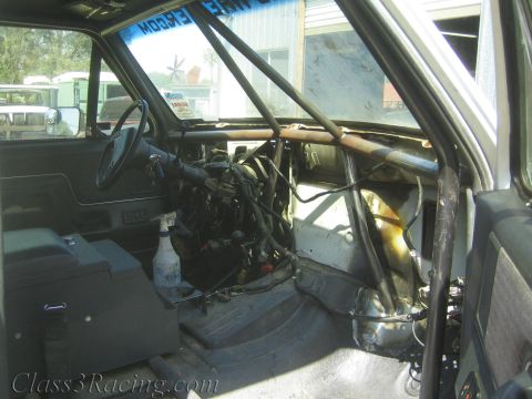 1990 Ford Bronco - View into a gutted off-road truck cab with steering wheel, roll cage and an exposed engine in a shop yard.