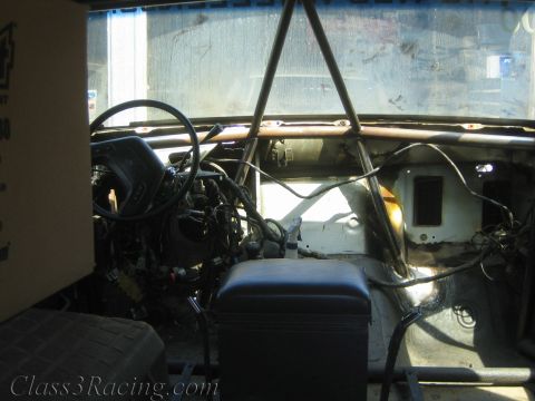 1990 Ford Bronco - Interior of a stripped truck showing a single seat, roll cage tubing and an exposed engine through the windshield.