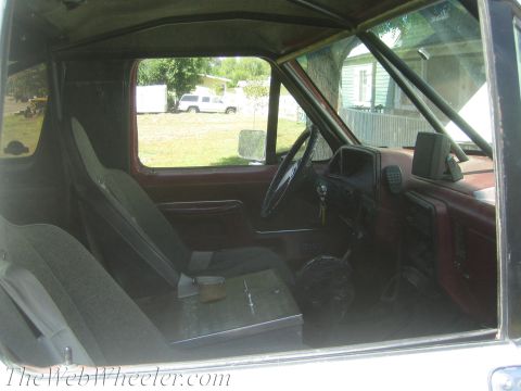 1990 Ford Bronco - View inside a worn truck cab with red door panels and dash, a fabric seat, floor shifter and a green yard visible through the side window.