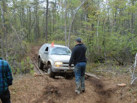 1991 Ford Bronco - A silver SUV drives through a wooded area on a dirt path, with a person standing nearby.
