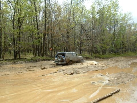 1991 Ford Bronco - A Ford Bronco navigates a challenging muddy off-road path surrounded by trees.