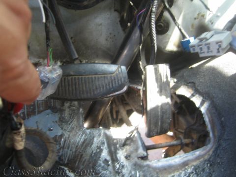 1990 Ford Bronco - grimy truck cab floor pan with a large cut-out hole near the brake and gas pedals, wiring and a hand visible at left.