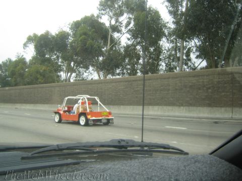 1990 Ford Bronco - small orange open-bodied utility-style car drives along a freeway beside a block sound wall, shot through a windshield.