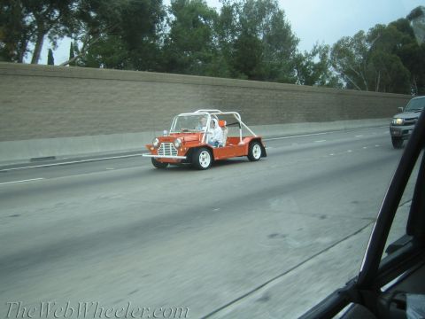 1990 Ford Bronco - orange open-top Mini-style buggy with a white roll structure carries a driver along a freeway past a sound wall.