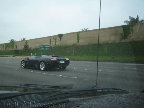 1990 Ford Bronco - black Lamborghini-style supercar cruises a freeway past a green 'San Clemente' exit sign and a tan retaining wall.
