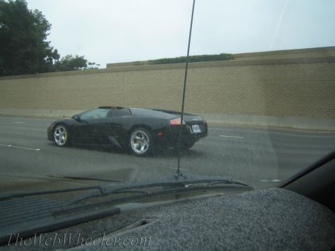 1990 Ford Bronco - black Lamborghini-style supercar travels a wet freeway, shot through a rain-streaked windshield with the wiper visible.