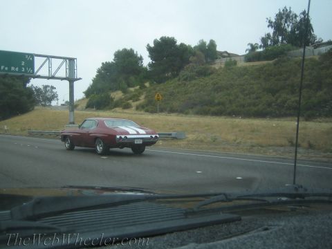 1990 Ford Bronco - dark red muscle car with twin white racing stripes drives a freeway past hillside homes, seen from a following car.