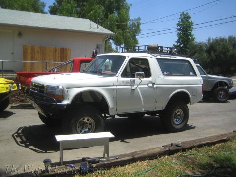 1990 Ford Bronco - lifted white full-size SUV with hardtop rides on knobby off-road tires in a driveway beside other project trucks.