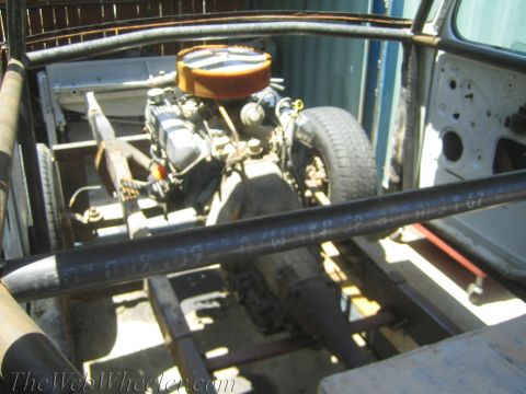 1990 Ford Bronco - engine with an orange round air cleaner sits on a stripped vehicle frame, viewed across tubular bars from the open cab area.
