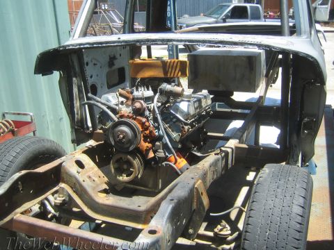1990 Ford Bronco - V8 engine with orange-painted accessories sits on a bare frame within a stripped, primer-gray vehicle body shell outdoors.