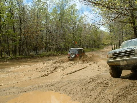 1991 Ford Bronco - Two off-road vehicles navigate a muddy trail surrounded by trees.