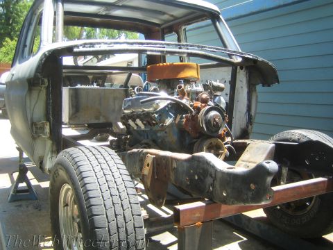 1990 Ford Bronco - V8 engine with an orange air cleaner sits on a frame inside a gutted vehicle cab shell beside a blue building.