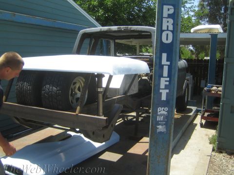 1990 Ford Bronco - man guides a white fiberglass front body clip down onto a tube-framed vehicle resting on a blue 'Pro Lift' hoist.