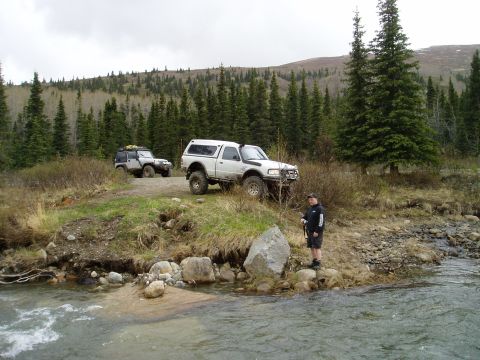 2000 Ford E350 - Two off-road vehicles are parked on a dirt path next to a flowing stream, with a man observing the water.